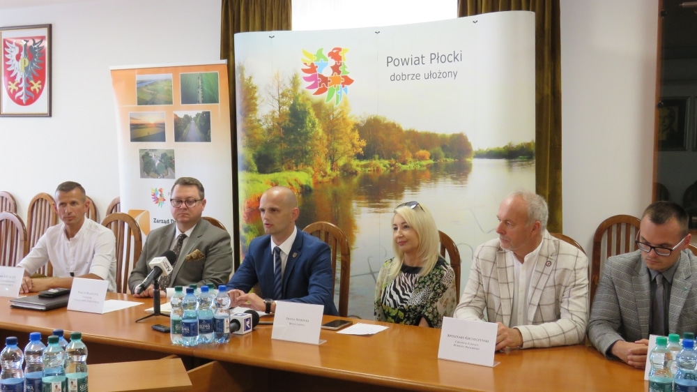 Konferencja prasowa w sali urzędowej Powiatu Płockiego, podczas której omawiane są plany przebudowy skrzyżowania w Wonicach, z obecnością lokalnych władz oraz przedstawicieli mediów.