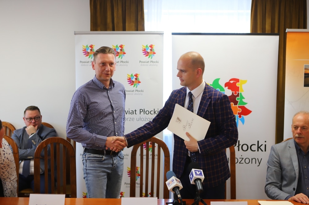 Na zdjęciu dwóch mężczyzn uścisnęło sobie dłonie podczas podpisywania umowy w biurze Powiatu Płockiego, w otoczeniu kilku innych osób.