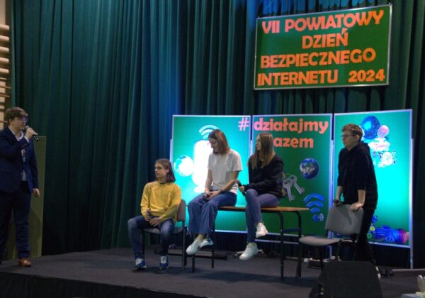 Uczestnicy VII Powiatowego Dnia Bezpiecznego Internetu w Gąbinie biorą udział w dyskusji na temat bezpieczeństwa w sieci, na scenie udekorowanej tematycznymi grafikami.