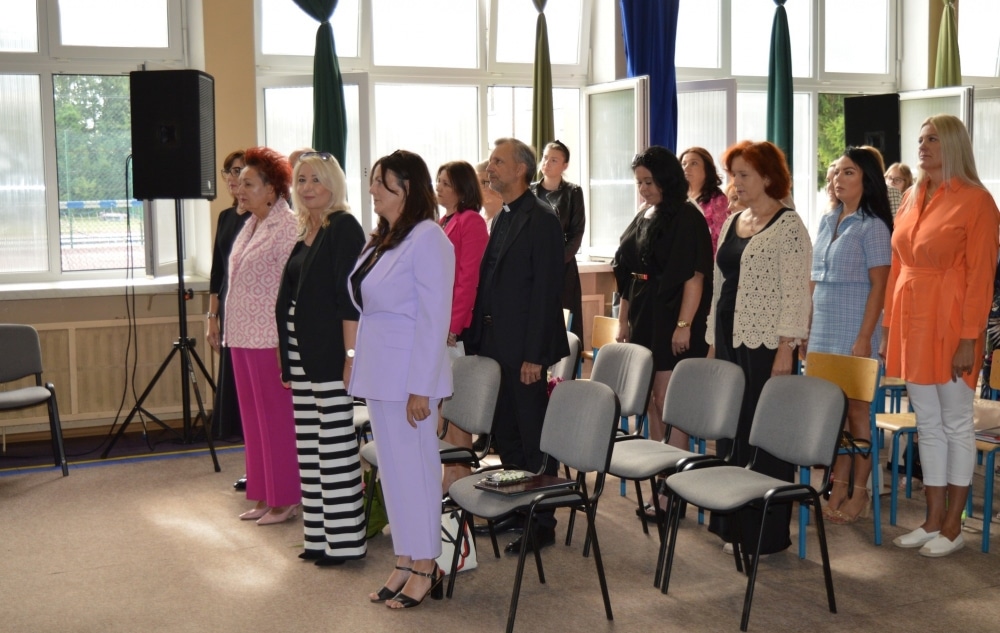 Uczestnicy uroczystości stoją w sali, ubrani w eleganckie stroje, podczas inauguracji nowego roku szkolnego.