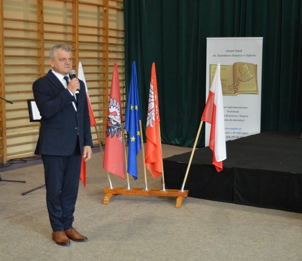 Na zdjęciu mężczyzna w garniturze przemawia do zgromadzonych, obok niego stoją flagi Polski, Unii Europejskiej oraz flagi lokalne.