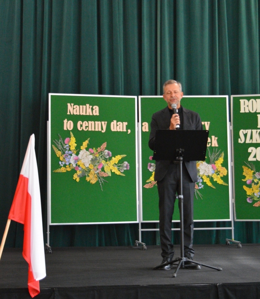 Mężczyzna w garniturze przemawia na scenie podczas uroczystości rozpoczęcia nowego roku szkolnego, obok znajdują się zielone tablice z napisami oraz polska flaga.