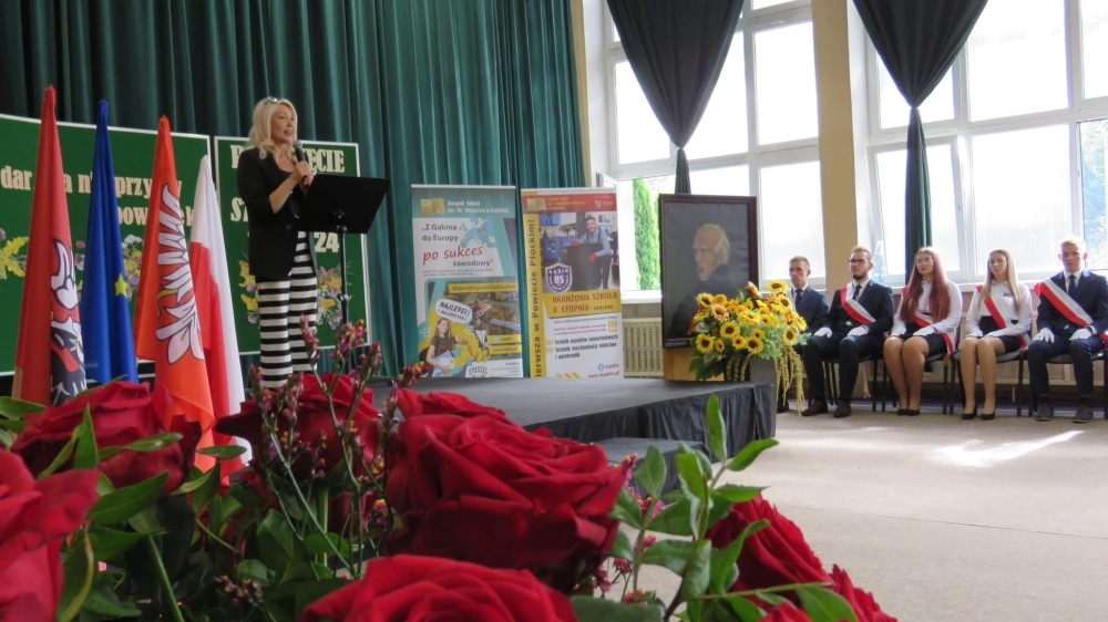 Na zdjęciu przedstawiona jest inauguracja nowego roku szkolnego w Zespole Szkół im. Stanisława Staszica, gdzie osoba mówiąca znajduje się na scenie, a w tle widoczne są flagi oraz uczniowie w strojach galowych.