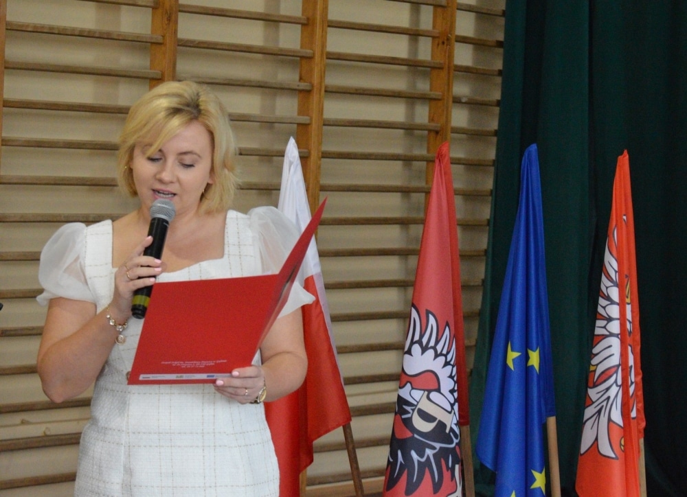 Na zdjęciu widać kobietę trzymającą mikrofon, która przemawia podczas uroczystości rozpoczęcia nowego roku szkolnego, obok flagi Polski oraz innych flag regionalnych.