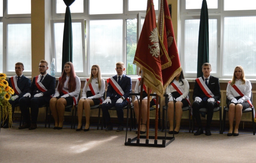Uczniowie w eleganckich strojach, siedzący w rzędzie, podczas ceremonii powitania nowego roku szkolnego, z flagami w tle.