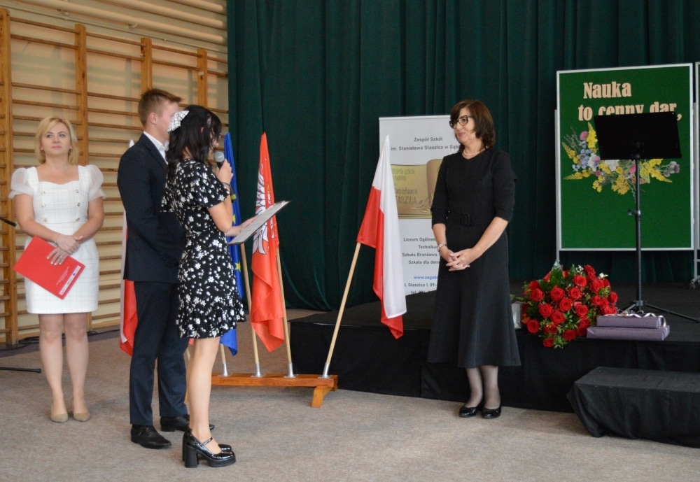 Uroczystość rozpoczęcia nowego roku szkolnego w zespole szkół, podczas której przemawia jedna z uczennic, a w tle widoczne są flagi i dekoracje.