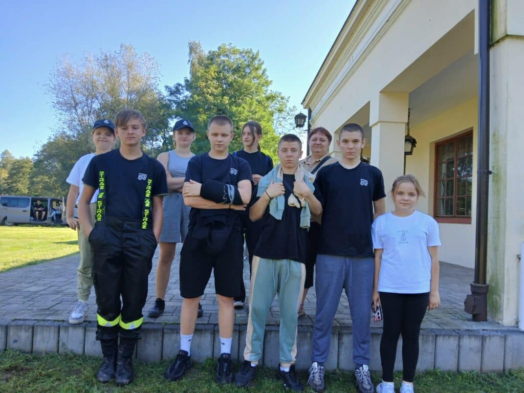 Na zdjęciu stoi grupa młodych ludzi w różnorodnych strojach, pozujących przed budynkiem podczas VI edycji spływu kajakowego w Cierszewie.