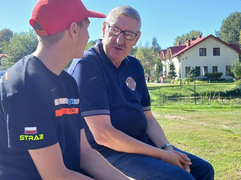 Na zdjęciu przedstawiono dwóch mężczyzn rozmawiających w słoneczny dzień w pobliżu wody, podczas VI edycji spływu kajakowego w Cierszewie.