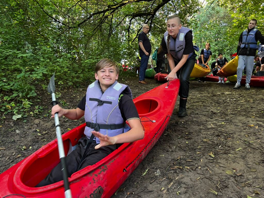 Uczestnicy VI edycji spływu kajakowego w Cierszewie przygotowują się do wodnej przygody, z uśmiechami na twarzach i w kajakach na brzegu.