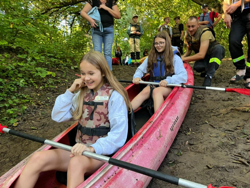 Dwie dziewczynki w kamizelkach ratunkowych przygotowują się do spływu kajakowego, uśmiechając się na tle leśnego krajobrazu.