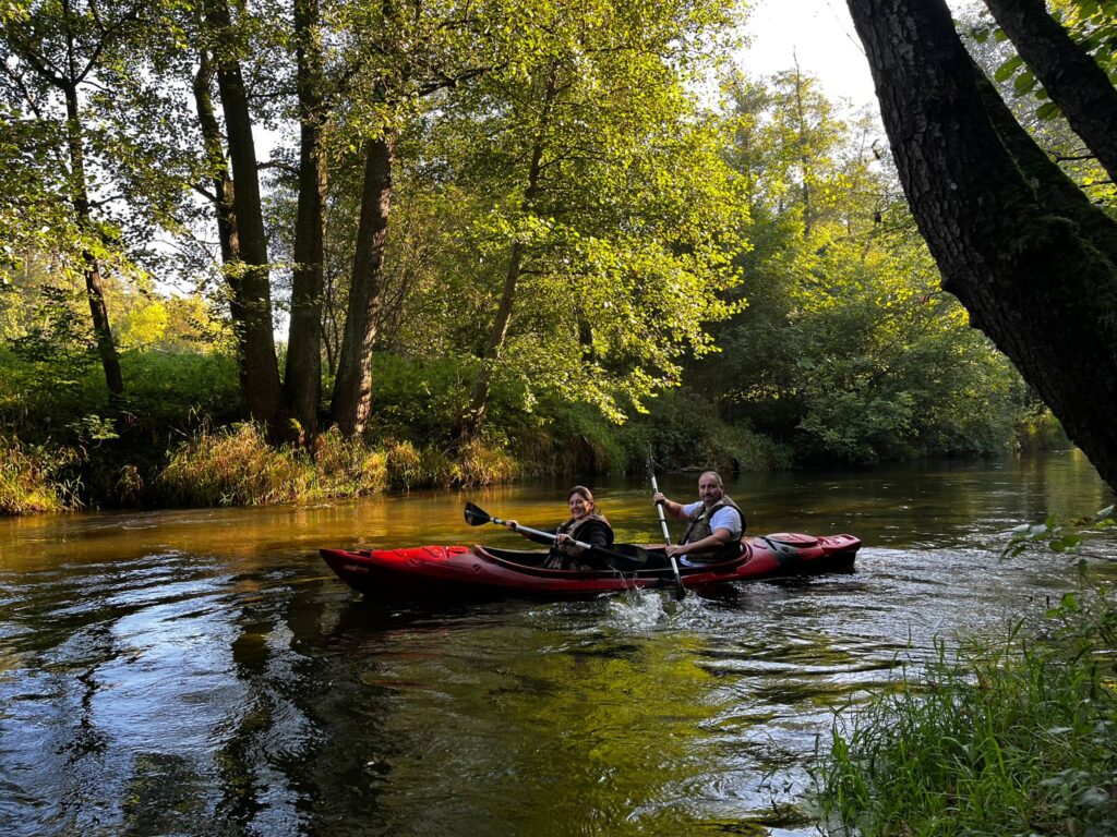 Dwoje uczestników kajakowego spływu na rzece otoczonej zielenią, uchwyconych w przyjemnej, słonecznej scenerii.