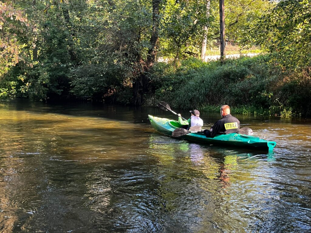 Na zdjęciu widać dwóch kajakarzy płynących rzeka w zielonym kajaku podczas VI edycji spływu kajakowego w Cierszewie, otoczonych przez bujną roślinność.
