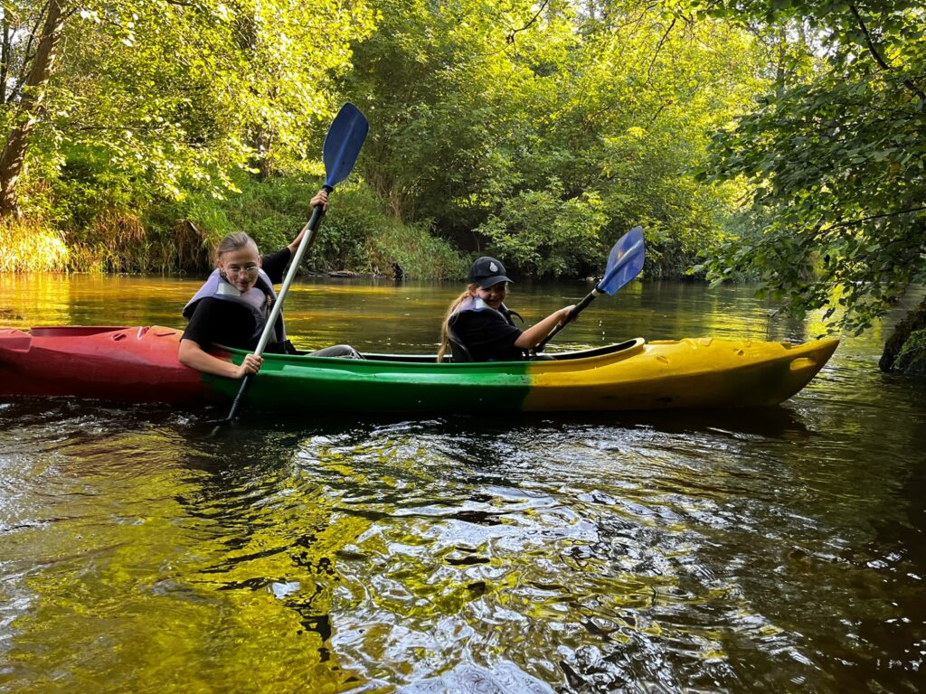Na zdjęciu widoczni są uczestnicy spływu kajakowego w Cierszewie, pływający po spokojnej rzece w otoczeniu bujnej zieleni.