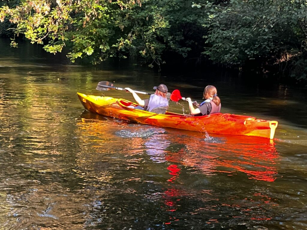 Na zdjęciu widać dwie osoby płynące kajakiem po spokojnej wodzie otoczonej zielonymi drzewami podczas VI edycji spływu kajakowego w Cierszewie.