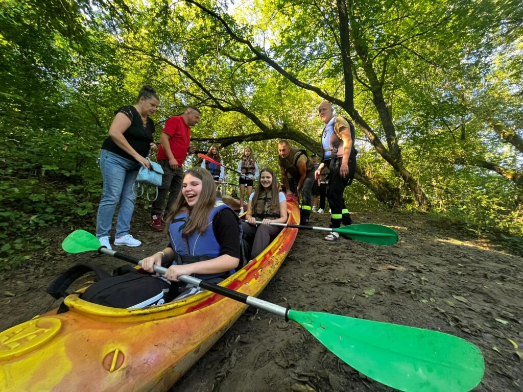 Uczestnicy VI edycji spływu kajakowego w Cierszewie przygotowują się do wodnej przygody, radośnie uśmiechając się i siedząc w kajaku na brzegu w otoczeniu zieleni.