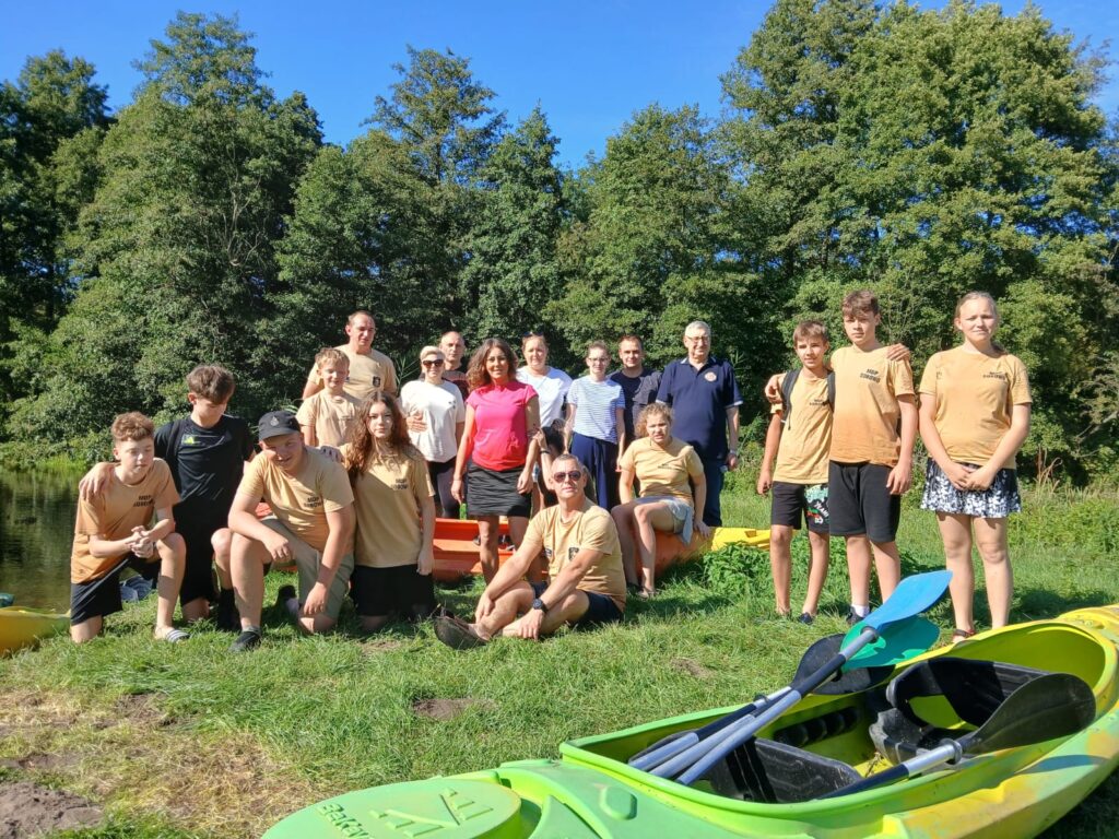 Grupa uczestników VI edycji spływu kajakowego w Cierszewie stoi na trawie obok kajaków, w otoczeniu zieleni i błękitnego nieba.