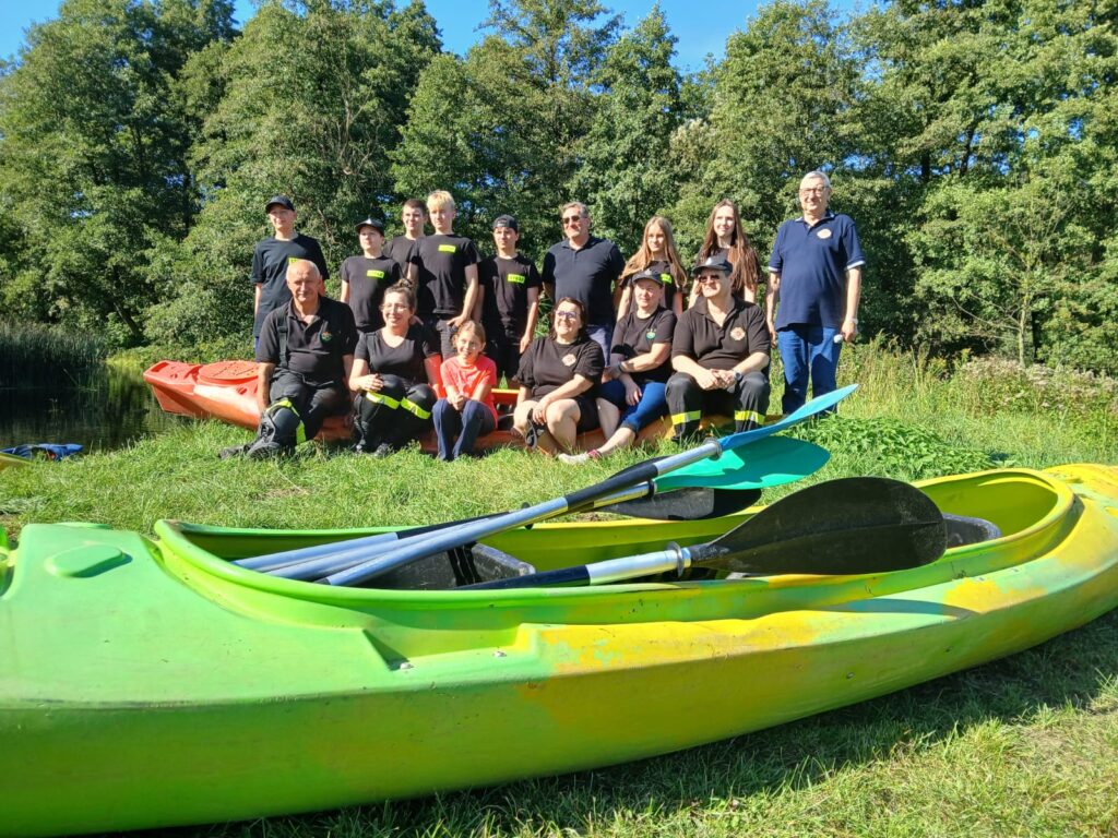 Grupa uczestników VI edycji spływu kajakowego w Cierszewie pozująca na tle kajaków i zielonej scenerii.