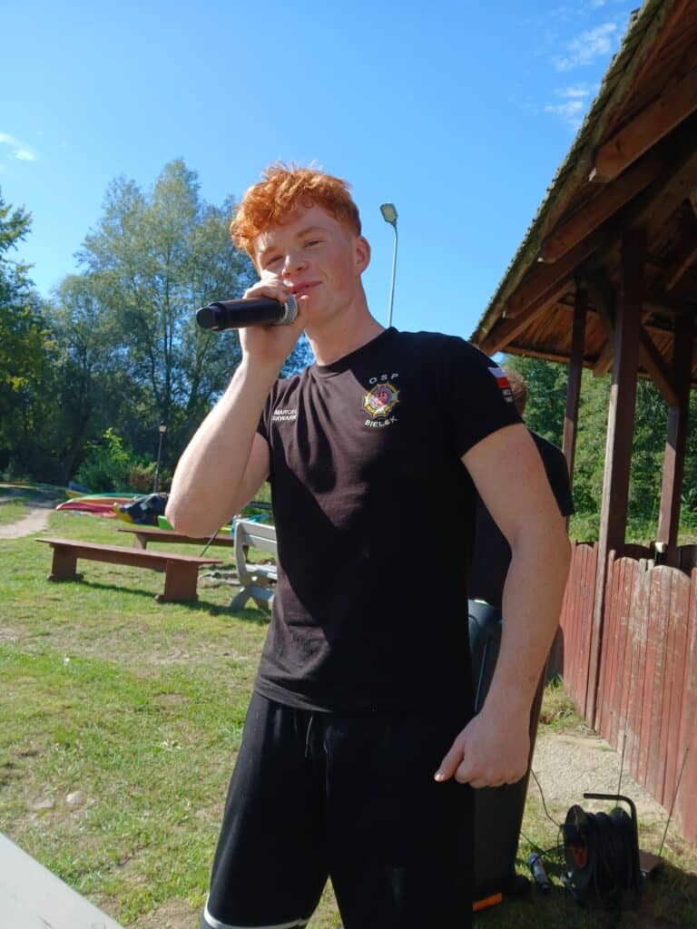 Mężczyzna z rudymi włosami śpiewa do mikrofonu podczas VI edycji spływu kajakowego w Cierszewie, w otoczeniu zieleni i błękitnego nieba.