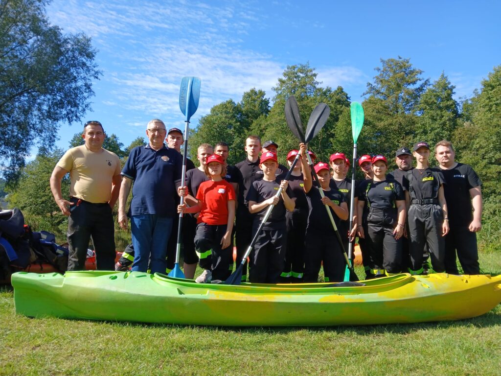 Uczestnicy VI edycji spływu kajakowego w Cierszewie pozują do zdjęcia w grupie, trzymając wiosła i stojąc obok kajaków.