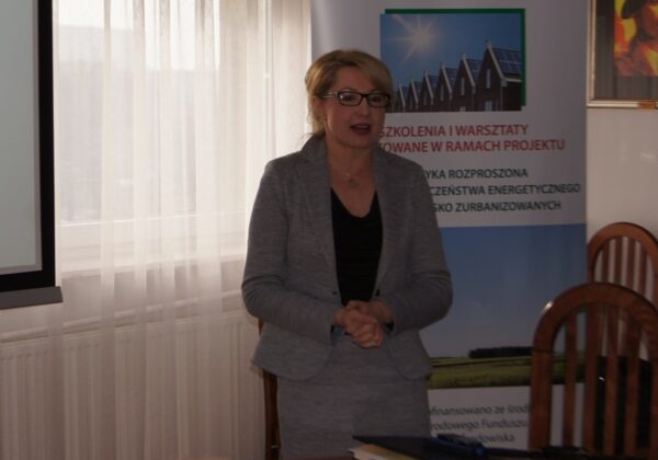 Na zdjęciu widoczna jest kobieta w szarym garniturze, która prowadzi prezentację na temat energetyki rozproszonej w kontekście bezpieczeństwa energetycznego na terenach nisko zurbanizowanych.