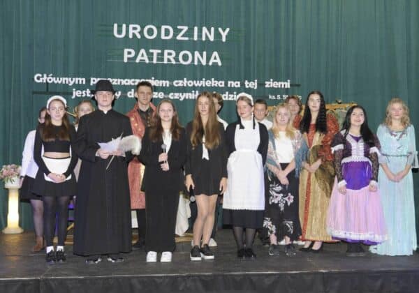 Grupa młodych ludzi w strojach historycznych prezentuje się na scenie z okazji urodzin patrona, z napisem „Urodziny Patrona” w tle.