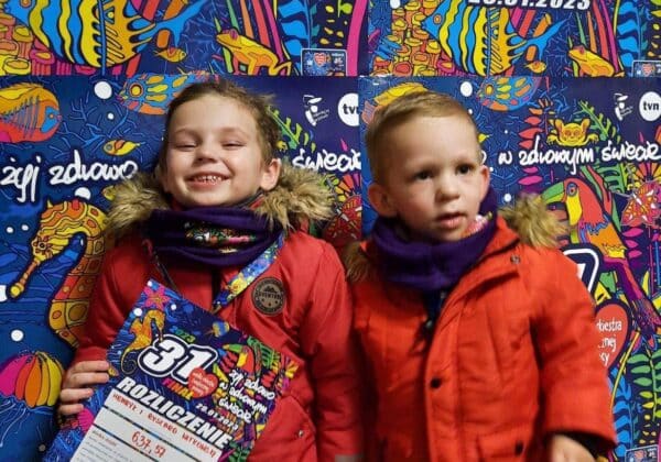 Dwoje dzieci stoi na tle kolorowego plakatu 31. Finału Wielkiej Orkiestry Świątecznej Pomocy, jedno z nich uśmiecha się szeroko, trzymając w ręku plakat z informacjami o wydarzeniu.