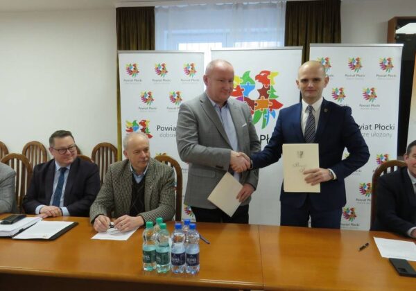 Na zdjęciu przedstawiono spotkanie, na którym dwóch mężczyzn uścisnął sobie dłonie po podpisaniu dokumentu, w otoczeniu innych uczestników oraz banerów promujących powiat płocki.