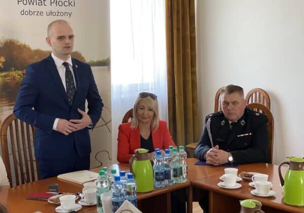 Na zdjęciu widać mężczyznę w garniturze przemawiającego do grupy osób, w tym kobietę w czerwonej marynarce oraz mężczyznę w mundurze, podczas spotkania w sali konferencyjnej.