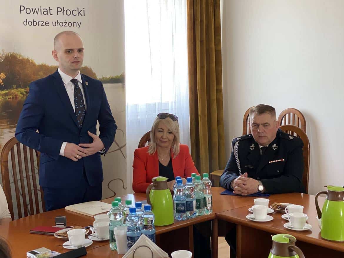 Na zdjęciu widać mężczyznę w garniturze przemawiającego do grupy osób, w tym kobietę w czerwonej marynarce oraz mężczyznę w mundurze, podczas spotkania w sali konferencyjnej.