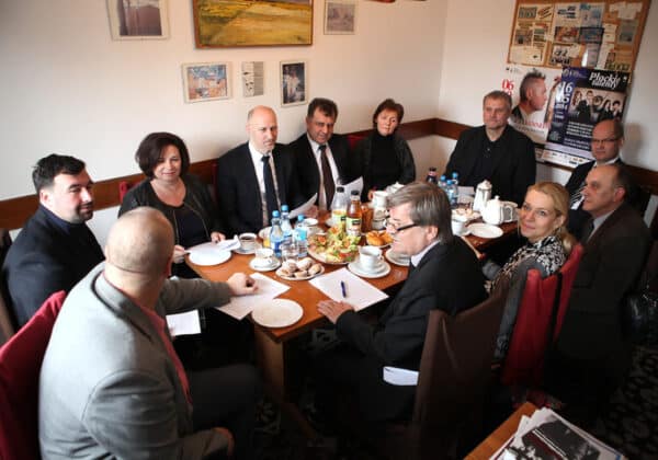 Na zdjęciu widoczna jest grupa osób siedzących przy stole, uczestniczących w spotkaniu, w otoczeniu kawy i przekąsek.