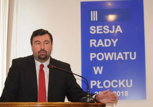 Mężczyzna w garniturze wygłasza przemówienie podczas sesji Rady Powiatu w Płocku, na tle niebieskiego plakatu informującego o sesji.