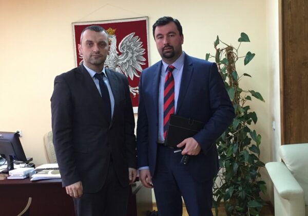 Na zdjęciu widać dwóch mężczyzn stojących w pomieszczeniu z roślinnością w tle, z herbem Polski na ścianie.