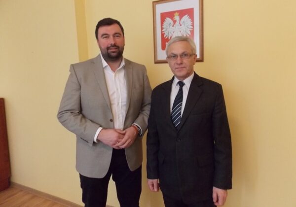 Na zdjęciu znajdują się dwaj mężczyźni, ubrani w eleganckie stroje, stojący obok siebie w pomieszczeniu z jasnymi ścianami i polskim godłem na tle.