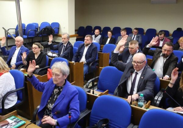 Na zdjęciu widać grupę osób siedzących w sali obrad, które podnoszą ręce w trakcie głosowania.
