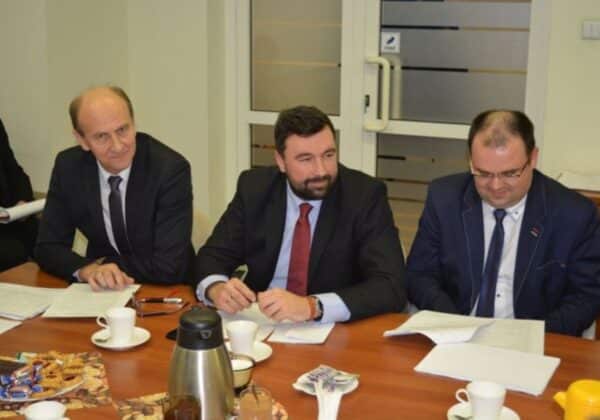 Na zdjęciu trzej mężczyźni w garniturach siedzą przy stole, uczestnicząc w spotkaniu.