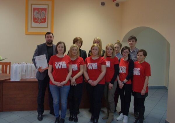 Grupa osób w czerwonych koszulkach z napisem „Super W” pozuje do zdjęcia w biurze, trzymając certyfikat i otoczeni przez białe torby.