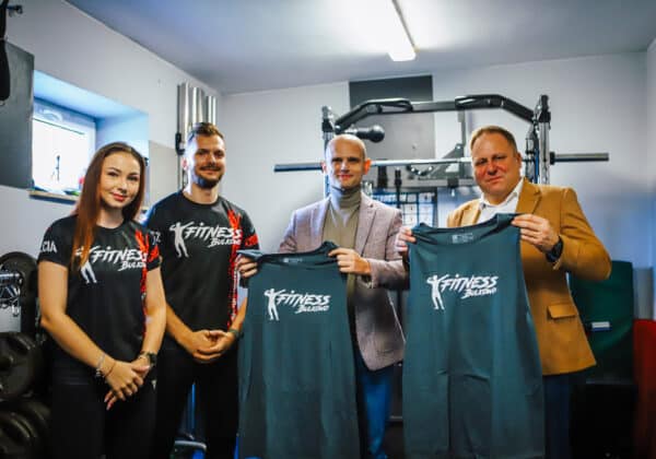 Czterech uczestników uśmiecha się, trzymając koszulki z logo "Fitness Bulkowo" w nowoczesnej siłowni.
