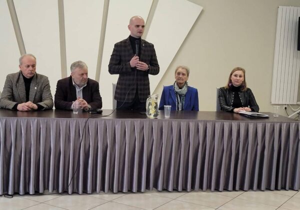 Na zdjęciu przedstawiono grupę osób siedzących przy stole, z mężczyzną stojącym i przemawiającym, w kontekście spotkania lub konferencji.