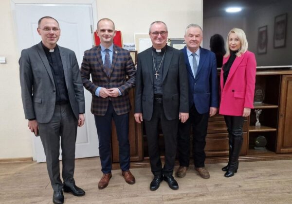 Na zdjęciu znajduje się grupa pięciu osób ubranych w eleganckie stroje, stojących w biurze z drewnianym meblem i telewizorem w tle.