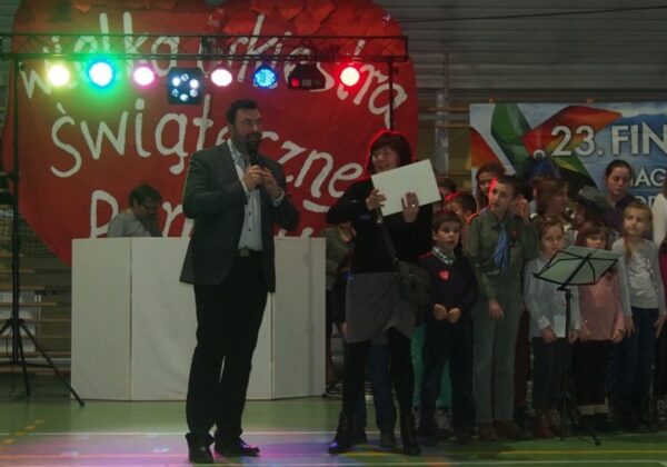 Na zdjęciu widać prowadzącego wydarzenie, który przemawia do zgromadzonej publiczności oraz grupę dzieci, na tle dekoracji związanej z 23. finałem WOŚP w powiecie płockim.