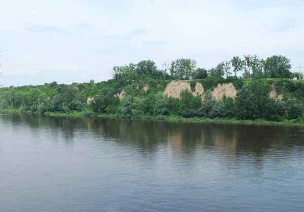 Widok na spokojną rzekę otoczoną zielonymi brzegami i piaszczystymi klifami na tle błękitnego nieba.