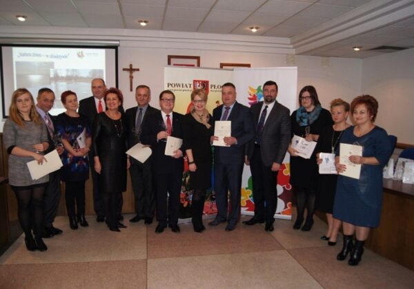 Na zdjęciu grupa osób stoi w sali, trzymając dyplomy i nagrody podczas uroczystości wręczenia „Kwiatów Powiatu”.