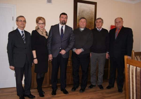 Na zdjęciu widocznych jest sześciu mężczyzn i jedna kobieta, stojących w eleganckich strojach w pomieszczeniu o jasnych ścianach i drewnianym wykończeniu.