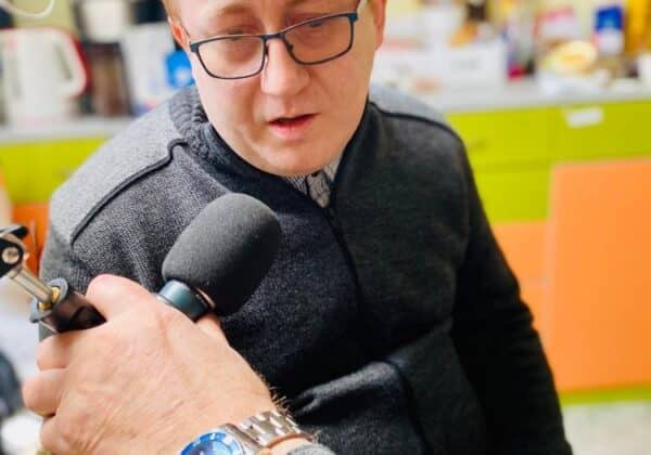 Mężczyzna w okularach siedzi w pomieszczeniu, podczas gdy ktoś z mikrofonem prowadzi z nim rozmowę.