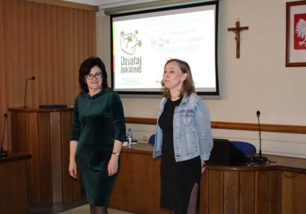 Na zdjęciu dwie kobiety stoją przy mównicy w sali, jedna z nich uśmiecha się, a w tle widać prezentację na temat projektu "Działaj lokalnie".