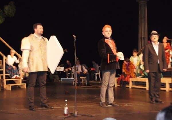 Na zdjęciu przedstawiona jest scena teatralna, na której mężczyźni w strojach kostiumowych prezentują się przed publicznością, przy akompaniamencie muzyków w tle.