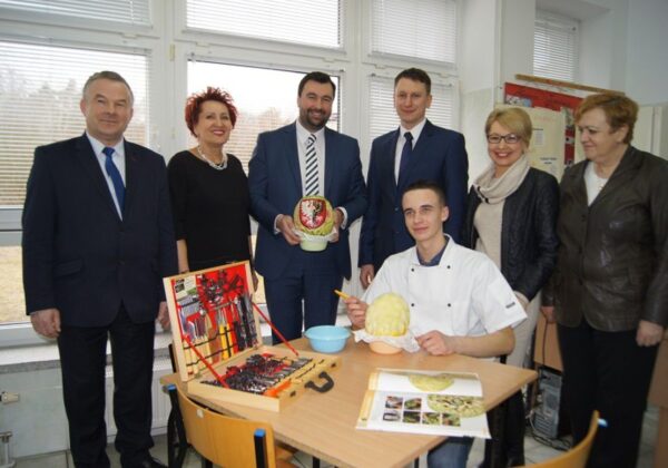 Na zdjęciu grupa osób w garniturach i w stroju kucharskim pozującą przy stole z przygotowanymi potrawami oraz narzędziami kuchennymi.