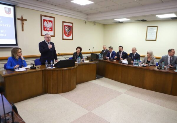 Na zdjęciu przedstawiono spotkanie członków rady powiatu, z mężczyzną przemawiającym przy mównicy oraz siedzącymi przy stole kobietami i mężczyznami.