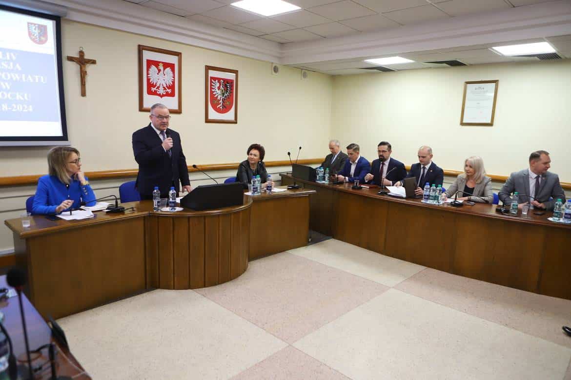 Na zdjęciu przedstawiono spotkanie członków rady powiatu, z mężczyzną przemawiającym przy mównicy oraz siedzącymi przy stole kobietami i mężczyznami.