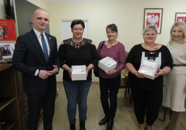 Na zdjęciu widać grupę pięciu osób, w tym mężczyznę w garniturze oraz cztery kobiety, które trzymają białe pudełka, w tle widoczne są flagi i dyplomy.